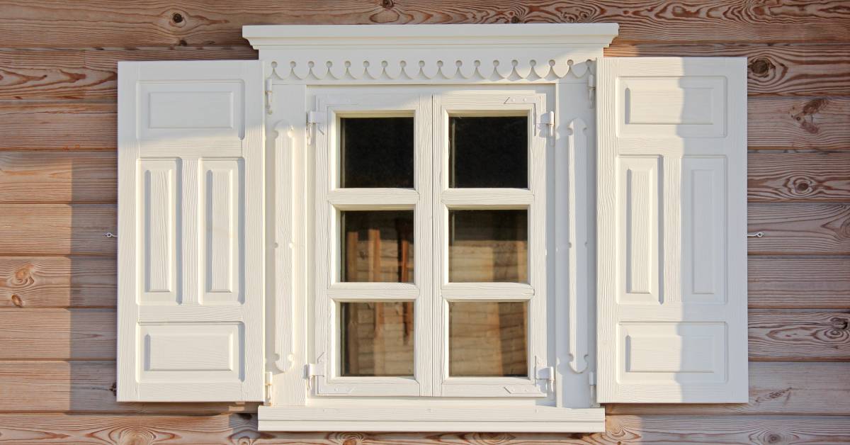 Closeup of a window on a light wooden house with intricate and stunning white wooden custom shutters.