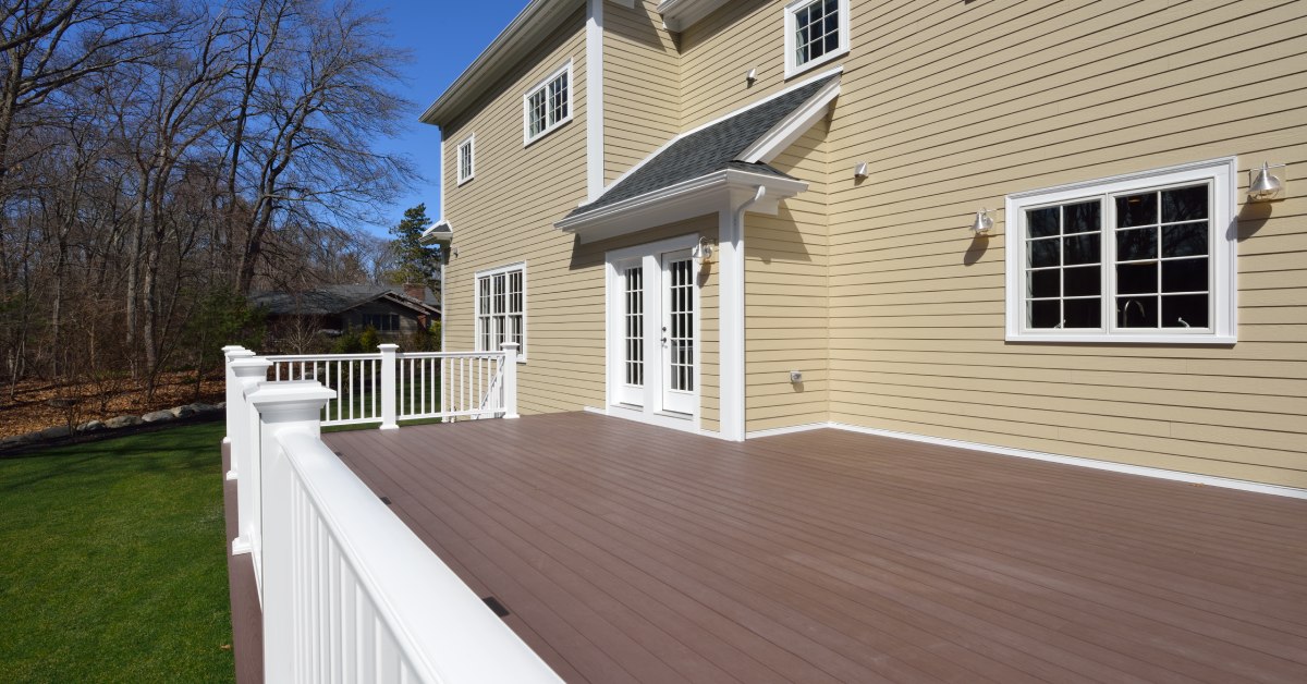 A large, empty deck in the backyard of a luxurious beige home with brown flooring and white wooden posts.